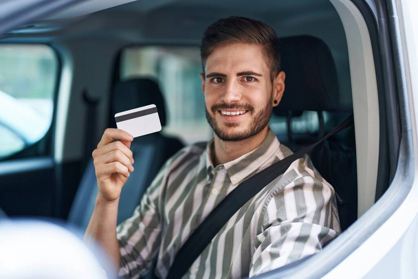 Hispanischer Mann mit Bart, der Auto fährt und Kreditkarte hält, sieht positiv und glücklich stehend aus und lächelt mit einem selbstbewussten Lächeln, das Zähne zeigt  - Foto, Bild