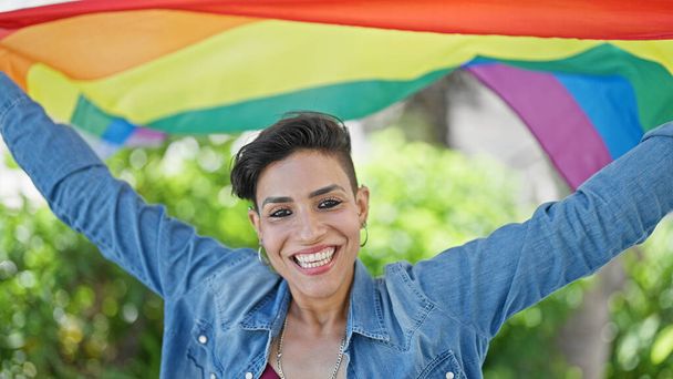 Giovane bella donna ispanica sorridente fiducioso tenendo bandiera arcobaleno al parco - Foto, immagini