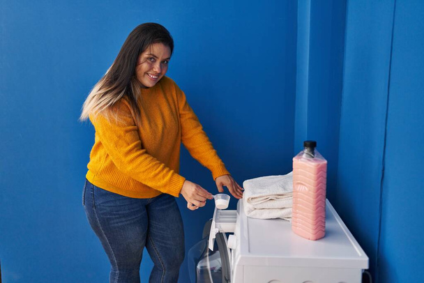 Молодая красивая плюс размер женщина улыбается уверенно выливая моющее средство на стиральную машину в прачечной - Фото, изображение