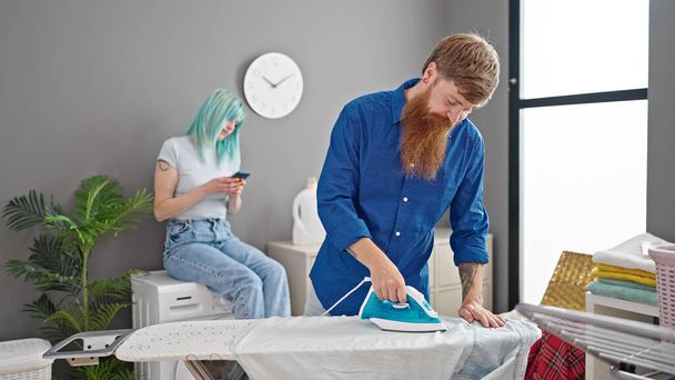 Man and woman couple ironing clothes while girlfriend use smartphone at laundry room - Photo, Image