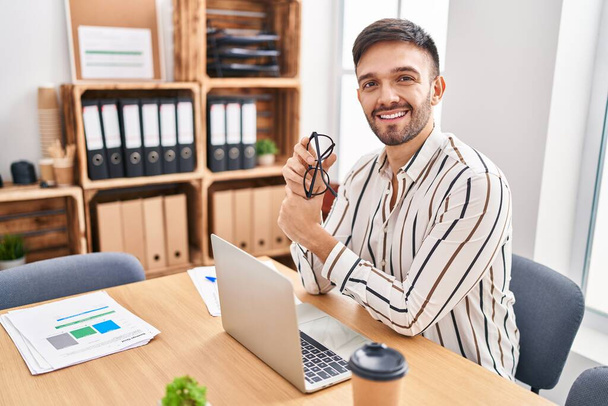 Young hispanic man business worker smiling confident sitting on table at office - Fotó, kép