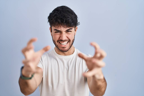 Hombre hispano con barba de pie sobre fondo blanco gritando frustrado por la rabia, manos tratando de estrangular, gritando loco  - Foto, imagen