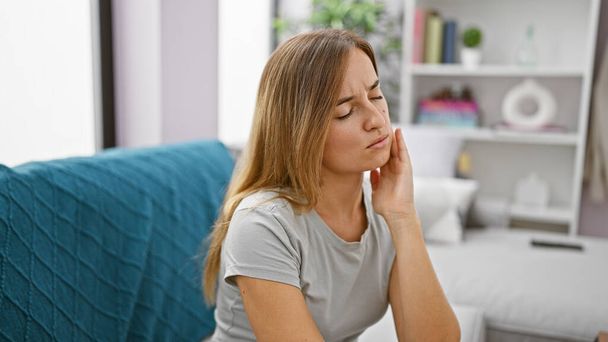 Vonzó fiatal szőke felnőtt nő ül a nappali kanapén otthon, szenved a kín súlyos fogfájás - orális kérdés igénylő fogorvos segítségét sürgősen. - Fotó, kép
