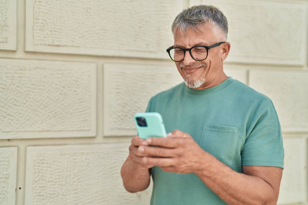 Middelbare leeftijd grijsharige man glimlachend vol vertrouwen met behulp van smartphone op straat - Foto, afbeelding