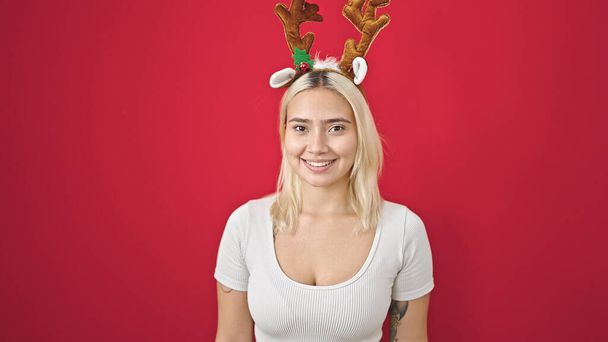 Young beautiful hispanic woman smiling confident wearing reindeer christmas hat over isolated red background - Foto, imagen