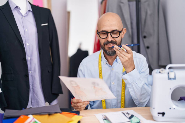 Young bald man tailor talking on smartphone looking clothing design at tailor shop - 写真・画像