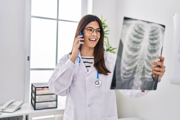 Young beautiful hispanic woman doctor talking on smartphone holding xray at clinic - Photo, Image