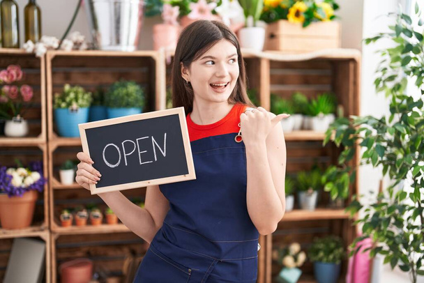 Mulher branca jovem trabalhando na florista segurando sinal aberto apontando polegar até o lado sorrindo feliz com a boca aberta  - Foto, Imagem