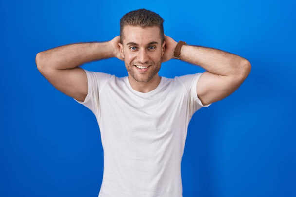 Junger kaukasischer Mann steht vor blauem Hintergrund und entspannt sich und dehnt sich, Arme und Hände hinter Kopf und Hals lächeln glücklich  - Foto, Bild