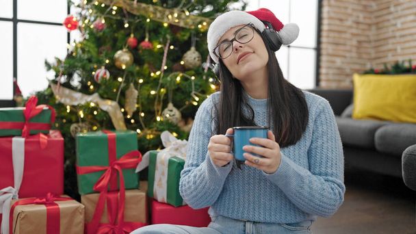 Jonge Spaanse vrouw die thuis naar muziek luistert met koptelefoon en kerstmuts op - Foto, afbeelding