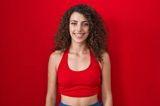 Ισπανίδα γυναίκα με σγουρά μαλλιά στέκεται πάνω από το κόκκινο φόντο με ένα χαρούμενο και δροσερό χαμόγελο στο πρόσωπο. τυχερό άτομο.  - Φωτογραφία, εικόνα