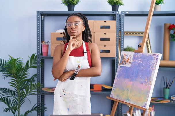 Junger Afroamerikaner mit Zöpfen im Kunstatelier Malerei auf Leinwand ernstes Gesichtsdenken über Frage mit der Hand am Kinn, nachdenklich über verwirrende Idee  - Foto, Bild
