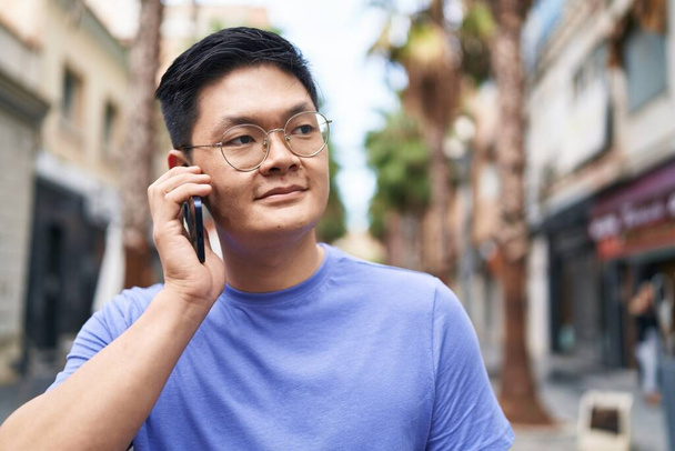 Genç Çinli adam gülümsüyor. Kendine güveni tam. Sokakta akıllı telefondan konuşuyor. - Fotoğraf, Görsel