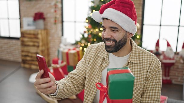 若いヒスパニックの男は自宅でクリスマスプレゼントを保持するスマートフォンでセルフィーを作ります - 写真・画像