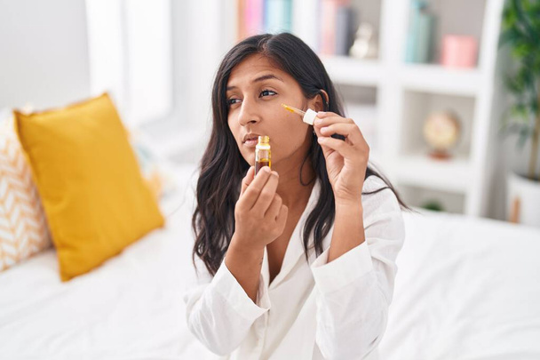 Young beautiful hispanic woman applying facial treatmet sitting on bed at bedroom - Foto, Imagem