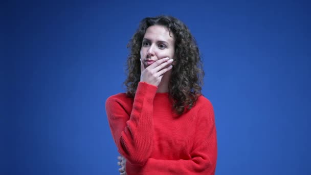 Töprengő nő, aki mélyen a kék háttérben áll piros pulóverben. 20-as évekbeli női személy átgondolt arckifejezéssel - Felvétel, videó