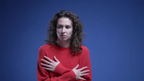 Verängstigte junge Frau umarmt sich zögernd und zeigt Angst und Panik vor blauem Hintergrund in rotem Pullover. Zwanziger hat Angst - Filmmaterial, Video