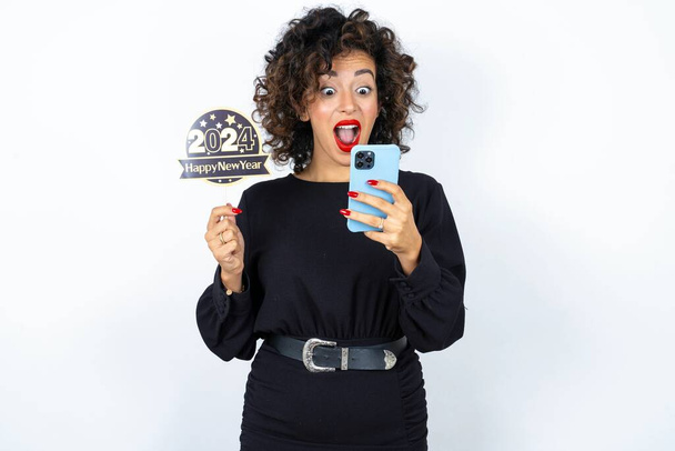 Siyah elbiseli, kıvırcık saçlı genç bir kadın elinde mutlu bir 2024 bayrağı, elinde telefonla para transferinden sonra şaşırmış hissediyor. - Fotoğraf, Görsel