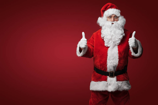 Porträt von isoliert auf grauem Weihnachtsmann mit erhobenem Daumen in rotem Anzug und Hut. - Foto, Bild