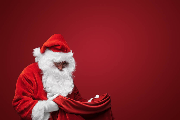 Noel Baba klasik kırmızı elbisesiyle, kendine güvenen bir çanta tutuyor. - Fotoğraf, Görsel