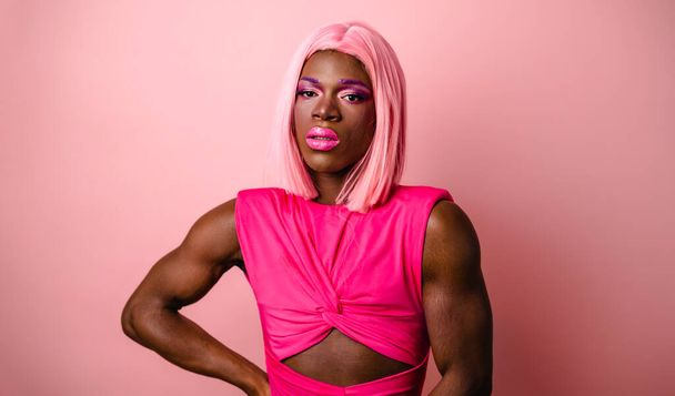 Mladý afroameričan drag queen nosit růžovou paruku a jasný make-up stojící s rukou v pase ve studiu při pohledu na kameru - Fotografie, Obrázek