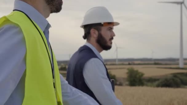 Інженери робітничої групи вітроелектростанції, що йдуть разом, планують млин у жовтому жилеті та шоломі, обладнані робочими інструментами на вітряній фермі. Професійні техніки чоловіки - Кадри, відео