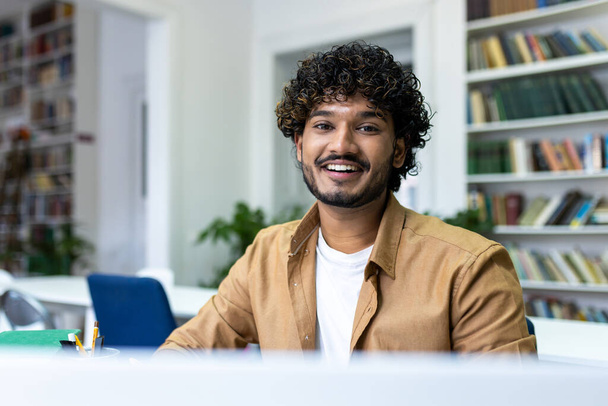 Κοντινό πλάνο πορτραίτο ενός νεαρού μουσουλμάνου φοιτητή που σπουδάζει σε ένα φορητό υπολογιστή σε μια πανεπιστημιακή βιβλιοθήκη, κοιτάζοντας και χαμογελώντας στην κάμερα. - Φωτογραφία, εικόνα