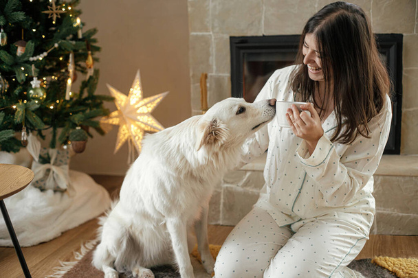 Bella donna in pigiama elegante con tè caldo rilassante con simpatico cane al camino accogliente, godendo mattina di Natale in soggiorno decorato festivo. Buon Natale! Vacanze invernali con animali domestici - Foto, immagini