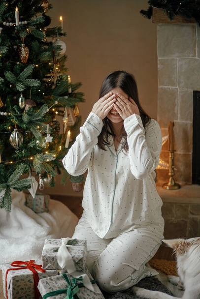 Šťastná žena ve stylovém pyžamu zavírající oči před stylovými vánočními dárky ve slavnostním zdobeném obývacím pokoji, užívající si vánoční ranní překvapení. Veselé Vánoce! Zimní dovolená - Fotografie, Obrázek
