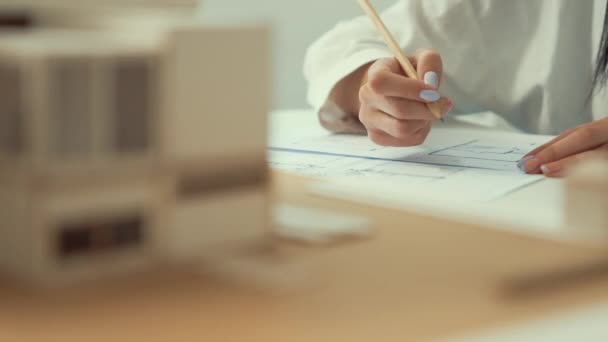 Detailní záběr na zkušené mladé krásné architekt inženýr ručně psaní dům konstrukce na návrh, zatímco ukazuje na dům model na pracovním stole. Rozmazané pozadí. Soustřeď se na ruce. Neposkvrněné. - Záběry, video
