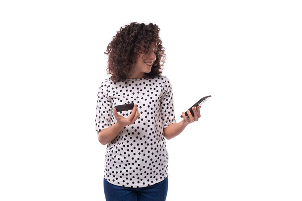 joven mujer caucásica de cabello oscuro vestida con una blusa muestra una tarjeta de plástico y un teléfono inteligente en sus manos. - Foto, Imagen