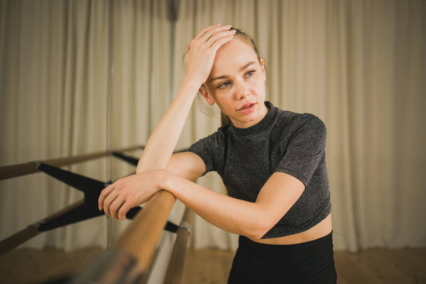 Upset ballerina in training suit stands near ballet barre and looks at the mirror in dance studio. Woman prepares for performance. Ballet dancer doing gymnastic exercises. Classical ballet school. - Photo, Image