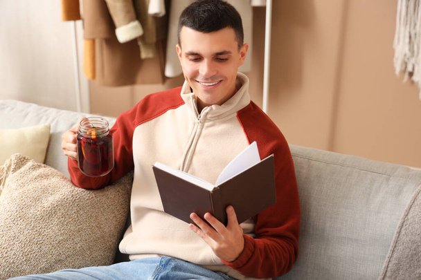 Przystojny młody człowiek ze szklanką ciepłego grzanego wina czyta książkę siedząc na szarej kanapie w salonie - Zdjęcie, obraz
