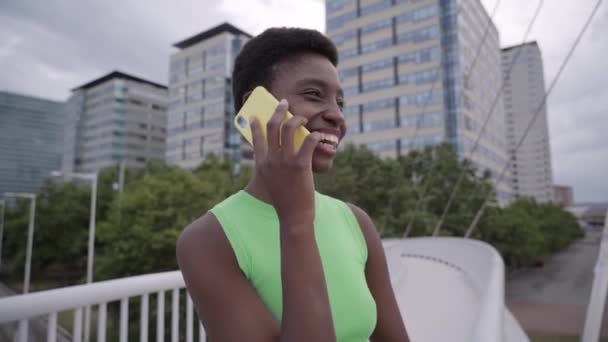 Gülümseyen Afro-Afrikalı kadın şehir merkezinde duran akıllı telefonu kullanarak sesli mesaj gönderiyor. Arka planda mobil hücre binaları olan güzel siyah kadın çağrısı, bağımlılık teknolojisi kavramı. - Video, Çekim