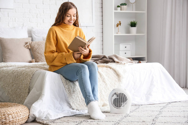 Mujer joven leyendo libro cerca de calentador de ventilador eléctrico en el dormitorio - Foto, imagen