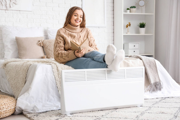 Fiatal nő könyvmelegítő lábakkal radiátoron a hálószobában - Fotó, kép