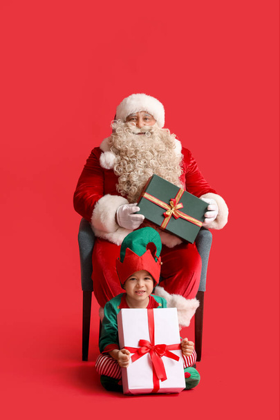 サンタクロースとかわいい小さなエルフ,赤い背景にクリスマスプレゼント - 写真・画像
