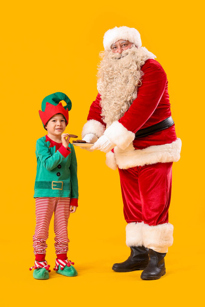 Santa Claus y lindo pequeño elfo comiendo galletas sobre fondo amarillo - Foto, imagen