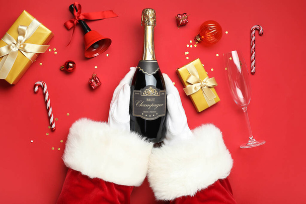 Manos de Santa Claus con botella de champán, decoraciones navideñas y regalos sobre fondo rojo, primer plano - Foto, imagen