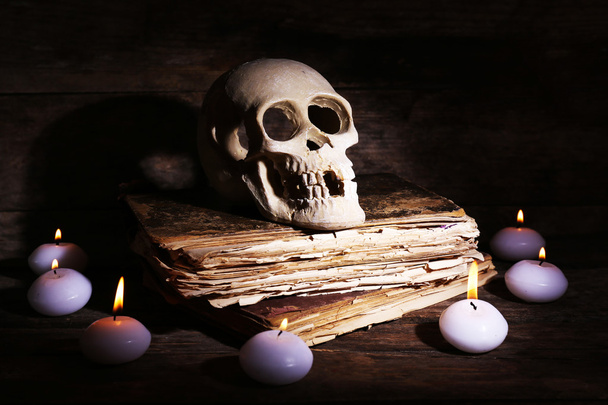 Still life with human skull, retro book and candlelight in dark on wooden table, closeup - 写真・画像