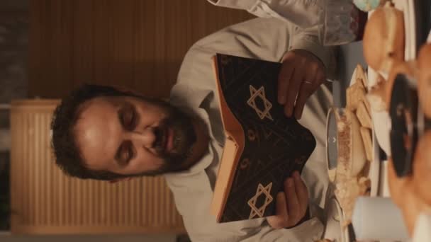 Medium vertical shot of middle-aged Jewish father with beard, in kippah sitting at festive table with traditional Hanukkah foods and reading aloud prayer from Torah, with star of David on cover - Footage, Video