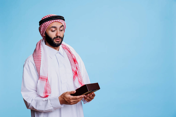 Ενθουσιασμένος μουσουλμάνος ντυμένος με παραδοσιακά ρούχα κρατώντας τυλιγμένο κουτί δώρου με προσμονή. Έκπληκτος arab πρόσωπο που λαμβάνει δώρο γενεθλίων, κοιτάζοντας giftbox με κορδέλα τόξο - Φωτογραφία, εικόνα