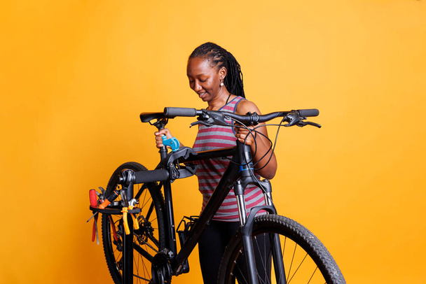 Gezond actief Afrikaans-Amerikaanse vrouw doet jaarlijkse fiets reparaties met behulp van gespecialiseerde werktuigen. Jeugdige zwarte vrouw grijpen een gebroken fiets te worden onderzocht en aangepast op een reparatie standaard. - Foto, afbeelding