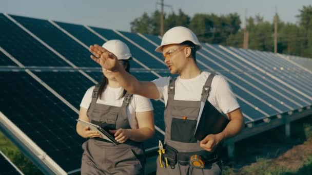 Két technikus, egy nő és egy férfi, napelemek sorai között sétálnak, és együtt beszélik meg egy napállomás működését. Zöld energia koncepció a naperőműben.Kiváló minőségű 4k felvételek - Felvétel, videó