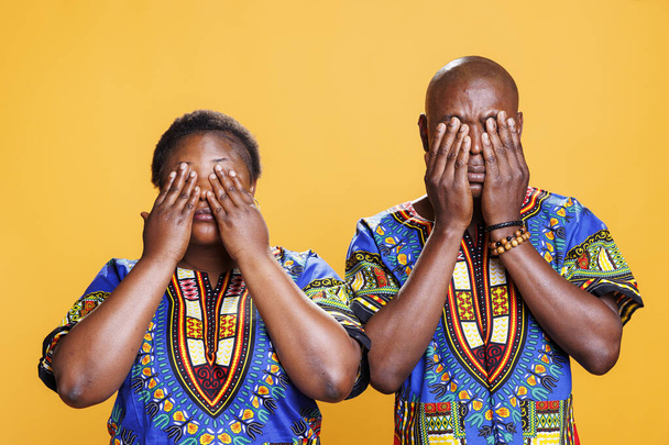 African american couple covering eyes with arms, showing hear no evil three wise monkeys portrait. Black man and woman pair holding palms on face on orange studio background - Photo, Image