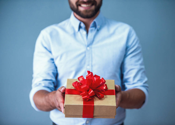 Обрезанное изображение красивого бородатого мужчины в элегантной повседневной одежде, держащего подарочную коробку и улыбающегося, на сером фоне
 - Фото, изображение