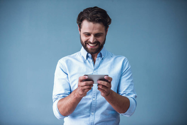 Knappe bebaarde zakenman in slimme vrijetijdskleding is met behulp van een slimme telefoon en glimlachend, op grijze achtergrond - Foto, afbeelding