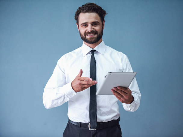 schöner bärtiger Geschäftsmann im formellen Anzug benutzt ein digitales Tablet, blickt in die Kamera und lächelt auf grauem Hintergrund - Foto, Bild