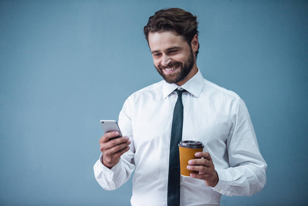 Yakışıklı sakallı işadamı resmi kıyafetli bir fincan kahve basılı tutup, gri arka plan üzerinde gülümseyen bir akıllı telefon kullanıyor - Fotoğraf, Görsel