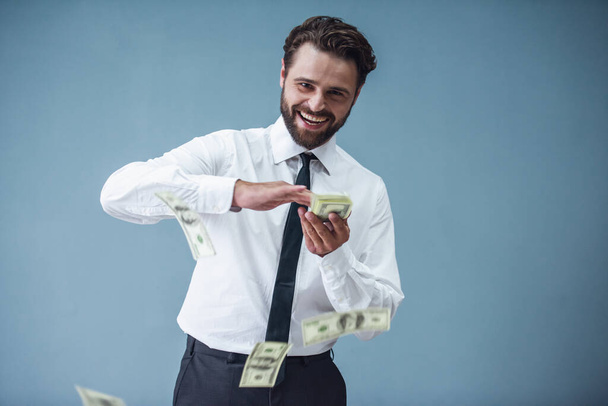 Όμορφος γενειοφόρος επιχειρηματία στο επίσημο κοστούμι ρίχνουν τα τραπεζογραμμάτια για και να χαμογελά, σε γκρίζο φόντο - Φωτογραφία, εικόνα